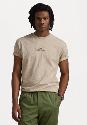 Bež pamukasta majica s roliranim rukavima i izvezenim tekstom "Polo Ralph Lauren". Uparena s hlačama s elastičnim pojasom u zelenoj boji. Ležeran kroj.