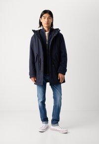 Navy blue parka with a zip front, drawstring waist, and large hood. Worn over a light sweater, paired with blue jeans and white sneakers.