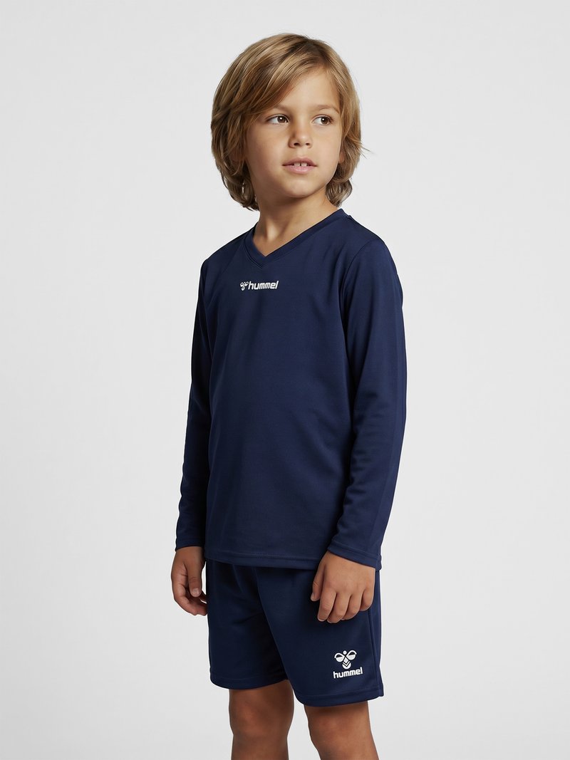 Jonge jongen die een marineblauw sportshirt met lange mouwen en bijpassende korte broek draagt, kijkt iets opzij tegen een effen achtergrond.