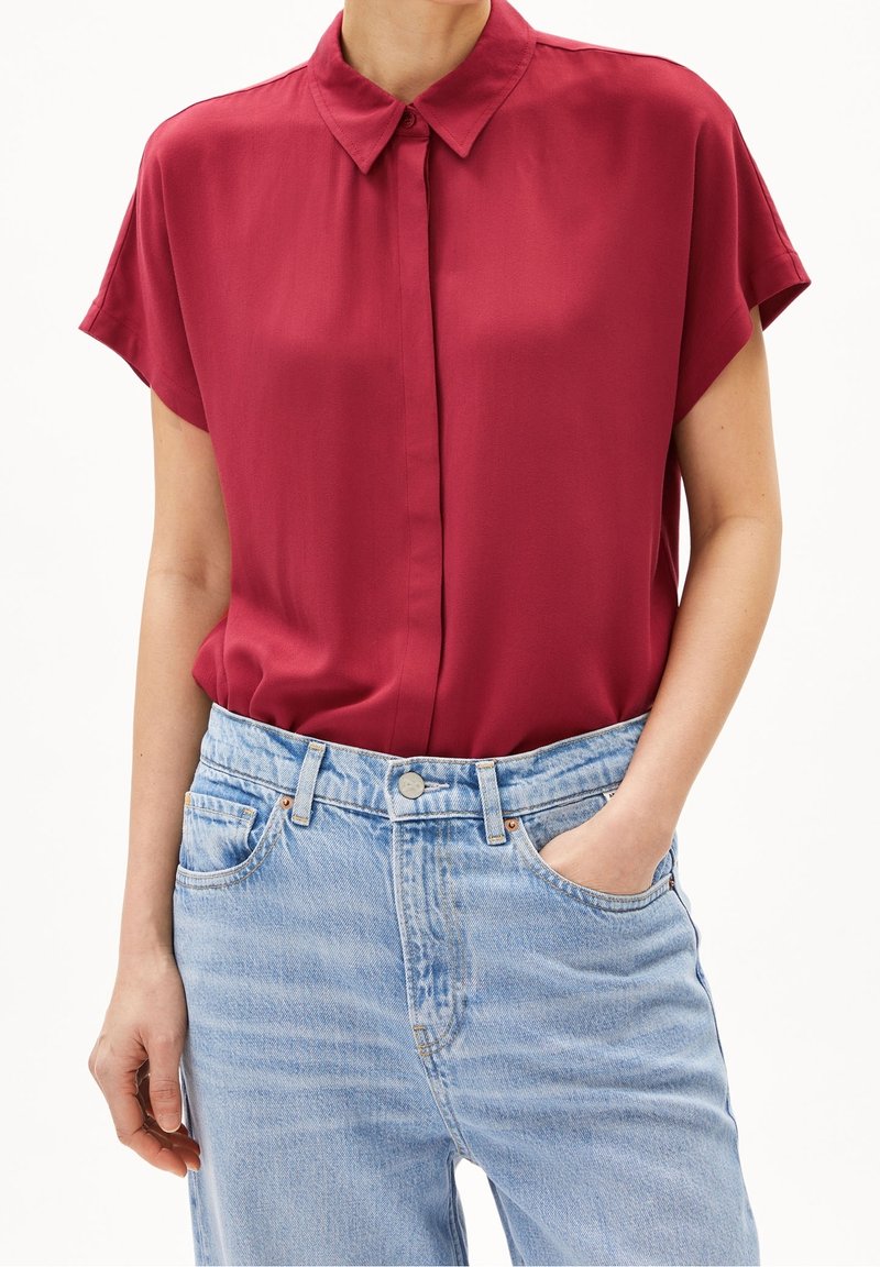 Femme portant un chemisier rouge à manches courtes et boutonné, rentré dans un jean bleu clair taille haute, avec une main dans une poche avant.