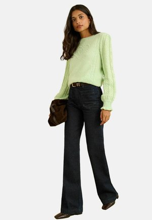 Helles grünes, kabelstrickartiges Sweatshirt mit Puffärmeln und Herzdesign. Kombiniert mit dunkelblauen, schmalen Schlagjeans und einer braunen Wildlederhandtasche.