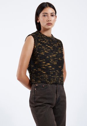 Kobieta o prostych ciemnych włosach, ubrana w czarny i żółty sweter bez rękawów oraz ciemne jeansy z wysokim stanem, stoi z rękami za plecami.