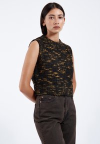 Stickad ärmlös topp i svart och guld med ett texturerat mönster, rund hals och kort silhuett, tillsammans med högmidjade mörka jeans.