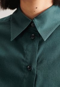 Mörkgrön skjorta med krage och en slät textur, med svarta knappar och en strukturerad krage. Nära vy av tyget och detaljerna.