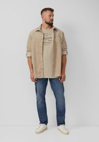 Beige corduroy overhemd over een lichtgrijs t-shirt met tekst, gecombineerd met blauwe jeans en witte sneakers. Eenvoudig ontwerp met contrasterende kleuren.