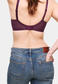 Violetter gepolsterter BH mit verstellbaren Trägern und Hakenverschluss, kombiniert mit mittelblauen Jeans mit mittlerer Taille, aufgesetzten Taschen und Zierstichen.