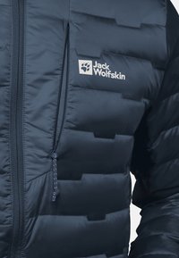Marineblaue Steppjacke mit gestepptem Design, durchgehendem Reißverschluss und verstellbarem Kordelzug. Jack Wolfskin-Logo in Weiß auf der Brust.