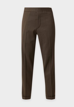 Hombre que lleva camisa beige de manga larga, pantalones verde oliva con bajos remangados, calcetines morados oscuros y zapatos de cuero marrón, de pie sobre fondo blanco.