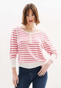 Raido pitkähihaine paita vaaleanpunaisessa ja valkoisessa, laajalla kaula-aukolla, napituslistalla ja ribbikuvioiduilla hiansuille. Pehmeä kangas, rento istuvuus.