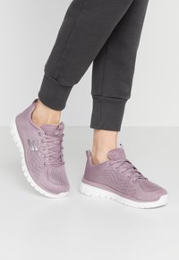 Lichtpaarse mesh atletiekschoenen hebben een gestructureerde bovenkant, een gevoerde tong en een witte rubberen zool met een zigzagpatroon.