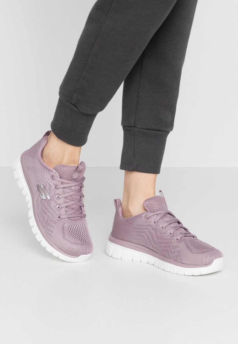 Lichtpaarse mesh atletiekschoenen hebben een gestructureerde bovenkant, een gevoerde tong en een witte rubberen zool met een zigzagpatroon.