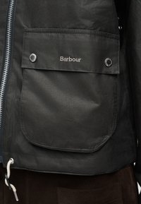 Poche de veste Barbour noire avec boutons-pression, logo brodé, fermeture éclair côté gauche et cordon blanc visible à l'ourlet inférieur.