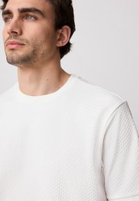 White textured short-sleeve shirt with a round neckline, featuring a subtle grid pattern and smooth fabric. Close-up view of the collar area.