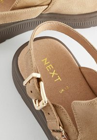 Sandales beige en daim doux avec une semelle texturée marron, comportant une boucle dorée et un logo en relief sur la semelle intérieure.
