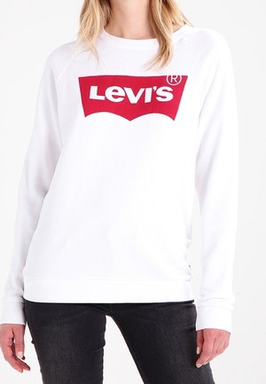 Osoba nosząca biały sweter z dużym czerwonym logo Levi’s na piersi, w połączeniu z czarnymi spodniami.