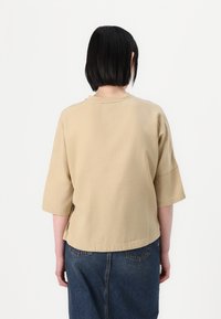 Beige cotton-blend top with short sleeves, crew neck, and a relaxed fit. The back features a straight hem and minimal seams.