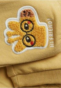 Gul plagg med en broderad handsdesign som har runda glasögon, röda kinder och texten "HEJ VÄN!" i vitt längs kanten.