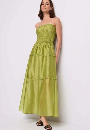 Donna in piedi contro il muro che indossa un abito lungo verde lime senza spalline con balze e sandali neri con punta aperta.