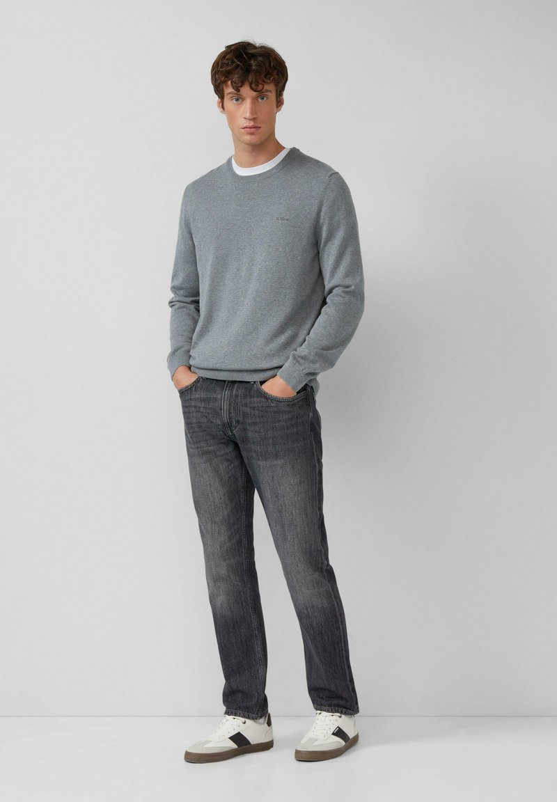 Man in een grijze ronde hals trui, donker gewassen jeans en witte sneakers met zwarte streep, staand met de handen in de zakken tegen een effen achtergrond.