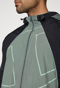 Ľahká bunda s zeleno-čiernou farebnou schémou, s geometrickým vzorom a predným zipsom. Materiál vyzerá štruktúrovaný a priedušný.