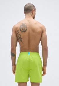 Pantalones cortos de baño de color verde brillante con una cintura elástica y una etiqueta azul. El diseño es simple, con una tela suave y ligera.