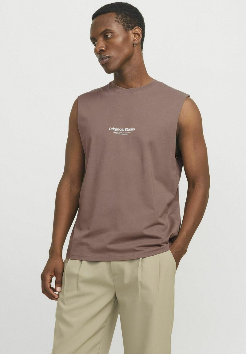 Camiseta sin mangas marrón con escote redondo, hecha de tela suave, con texto blanco en la parte frontal. Combinada con pantalones beige.
