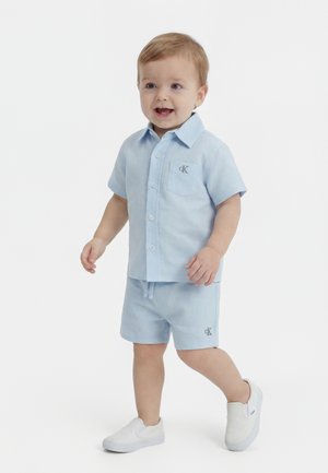 Toddler boy walking, wearing light blue short-sleeve button-up shirt and matching shorts with white slip-on shoes, smiling.