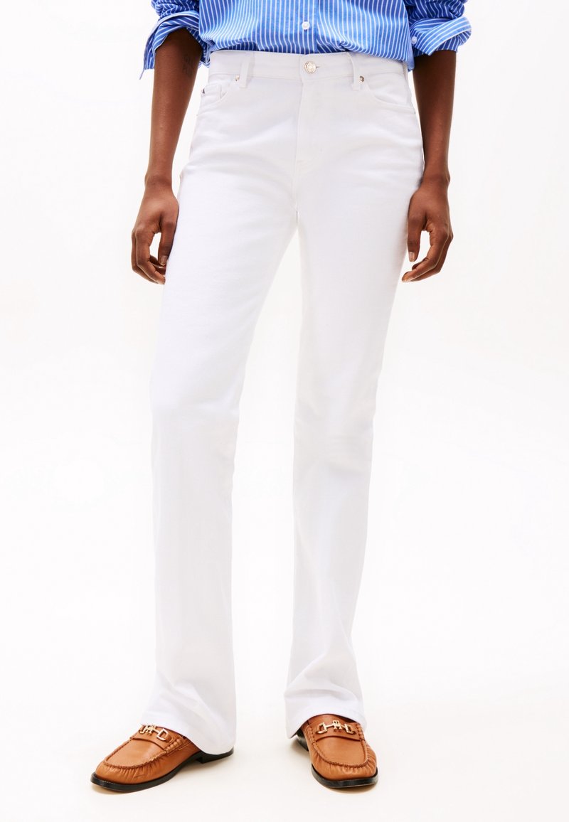 Personne portant un pantalon blanc à jambes droites, une chemise rayée bleue avec les manches retroussées, et des mocassins bruns avec un détail métallique.
