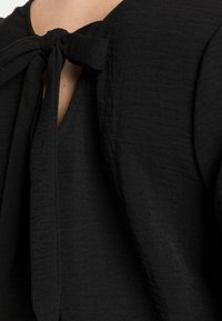 Blusa nera con tessuto testurizzato, apertura a chiave sul retro e dettaglio fiocco annodato al colletto.