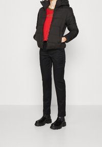 Doudoune noire avec capuche, associée à un t-shirt rouge, un pantalon cargo noir et des bottes à talons épais noires. Texture lisse et design ajusté.