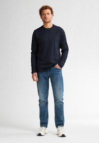 T-shirt a maniche lunghe blu navy con logo testurizzato, abbinato a jeans azzurri e sneakers bianche, caratterizzato da una vestibilità rilassata e scollo rotondo.