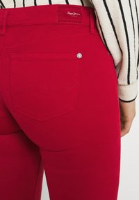 Primer plano de una persona que lleva puestas unas brillantes jeans rojos de Pepe, con un bolsillo trasero, combinados con una manga de suéter a rayas en crema y negro.