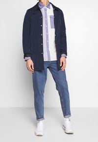 Chaqueta ligera de navy con botones a presión sobre una camisa de rayas multicolores. Jeans azules y zapatillas blancas completan el atuendo. Look casual y en capas.