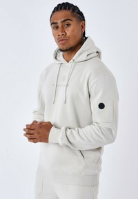 Sudadera gris claro con un bolsillo canguro, cordón ajustable y puños acanalados. Presenta un acento circular negro en la manga izquierda.
