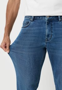 Sinimavad teksad, millel on veidi tuhm finish. Kanga materjalil on venivuse omadused ja taskud ning õmblusdetailid on selgelt nähtavad.