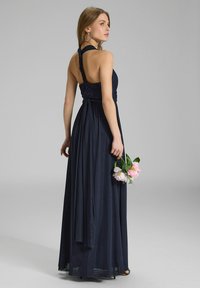 Lungo abito blu navy con scollo a halter intrecciato e gonna fluttuante. Schiena impreziosita da dettagli in pizzo, modello che tiene un bouquet di peonie.