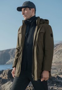 Oliwkowa parka z kapturem, z zamkiem z przodu i wieloma kieszeniami. Nosi się ją na czarną bluzkę z golfem, w połączeniu z czarnymi spodniami i szarym czapką.