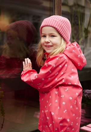 Kind in een felroze waterdichte jas met fruitpatronen en een bijpassende geribbelde roze muts, leunend tegen een raam.