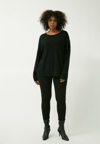 Schwarzer übergroßer Pullover mit langen Ärmeln, geripptem Saum und runder Ausschnitt, kombiniert mit schwarzen Leggings und knöchelhohen schwarzen Absatzstiefeln.
