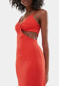 Červené šatové šaty s texturou, tenkými ramínky a detailem uzlu na poprsí. Obsahují výřezy po stranách a přiléhavý střih. Hladká látka.