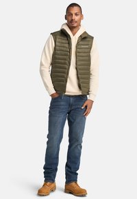 Groene pufferw vest over een crèmekleurige hoodie, gecombineerd met blauwe jeans en tan kleurige werklaarzen. Het vest heeft horizontale quiltpatronen en een ritssluiting aan de voorkant.
