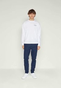 Chemise blanche à manches longues avec logo sur la poitrine gauche, associée à un pantalon bleu marine et des baskets blanches. Design simple et décontracté avec une coupe ample.