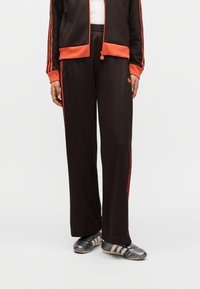 Καφέ αθλητική φόρμα με πορτοκαλί ρίγες, που περιλαμβάνει μπουφάν με φερμουάρ και φαρδιά παντελόνια. Συνοδεύεται από μαύρα και ασημί αθλητικά παπούτσια.