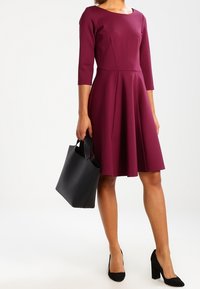 Nainen, jolla on polven pituinen burgundinvärinen mekko, jossa on kolmannes pitkähiha, pitää mustaa käsilaukku ja käyttää mustia paksupohjaisia kenkiä.