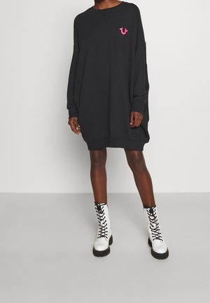 Person iført en oversized sort sweatshirt-kjole med et lille pink logo og hvide snørestøvler står på en ensfarvet baggrund.