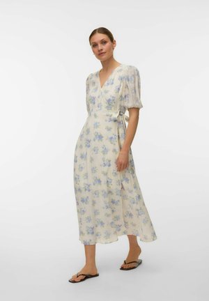 Femme portant une robe portefeuille midi blanche à imprimé floral bleu, manches ballon et sandales plates noires, debout devant un fond uni.