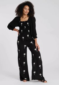 Mono negro con bordado de palmeras, tejido texturizado, escote cuadrado, mangas abullonadas y diseño de pierna ancha. Se llevan sandalias planas.