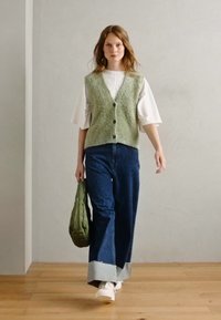 Groene gebreide vest met knopen, witte top met wijde mouwen, donkerblauwe jeans en grote manchetten; model houdt een groene tas vast, witte sneakers.