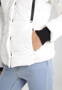 Bílá péřová bunda s prošívaným designem, černými žebrovanými manžetami a kapsami na zip. Tkanina vypadá hladce a je voděodolná.
