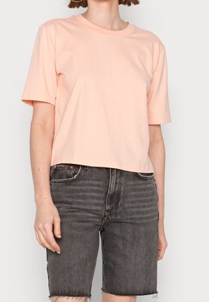 Osoba ubrana w jasno brzoskwiniowy t-shirt z krótkim rękawem i czarne, wysokiej talii szorty dżinsowe z postrzępionymi brzegami na tle jednolitym.